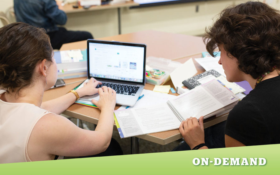Two educators sitting at table looking at a laptop and textbook.