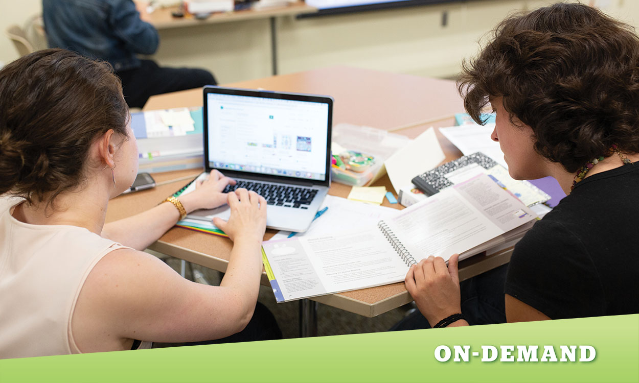 Two educators sitting at table looking at a laptop and textbook.