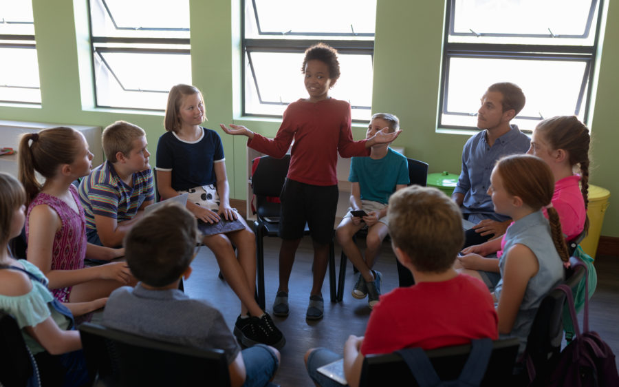 Front view of a diverse group of elementary school kids sitting on chairs in a circle and interacting during a lesson, one African American girl standing and talking while her classmates and male Caucasian teacher sit and listen